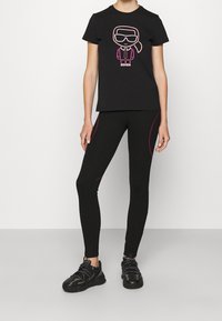 Camiseta negra con un diseño gráfico en rosa y blanco de un personaje con gafas de sol. Leggings negros con líneas de acento en rosa y zapatillas negras.