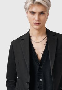 Svart blazer över en svart, sliten stickad cardigan med djup V-ringning. Har ett silverkedjearmband och synliga örhängen.