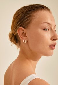 Silver hoop earrings with a twisted design, hanging close to the ear. The model has straight, slicked-back hair and a smooth complexion.