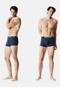 Boxers bleu marine avec une taille noire arborant le logo Pierre Cardin ; présentés sur un mannequin masculin sous plusieurs angles, pieds nus.
