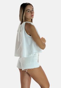 Witte mouwloze top met een losse pasvorm en strikdetail aan de achterkant, gecombineerd met witte denim shorts met rafelige zoom.