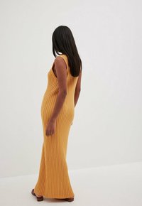 Robe longue côtelée de couleur moutarde ; design sans manches avec une silhouette ajustée, caractérisée par une texture lisse et un ourlet jusqu'au sol.