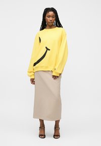 Femme aux longues tresses portant un sweat-shirt jaune oversize avec un smiley noir et une jupe midi beige avec des talons noirs.