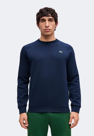 Man draagt een marineblauwe lange mouwen sweater met een klein groen krokodillenlogo op de borst en een groene broek, staand tegen een effen achtergrond.