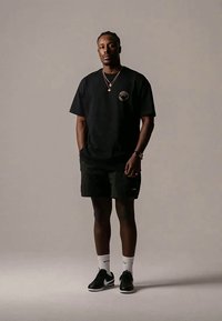 Junger Mann steht, trägt ein schwarzes T-Shirt mit kleinem Logo, schwarze Shorts, weiße Socken, schwarze Nike Sneakers, goldene Halsketten und eine Uhr vor einem neutralen Hintergrund.