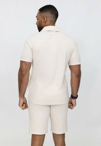 Ensemble de chemise à manches courtes et de shorts beige clair avec un motif en zigzag texturé. Le tissu semble lisse et décontracté, adapté aux temps chauds.