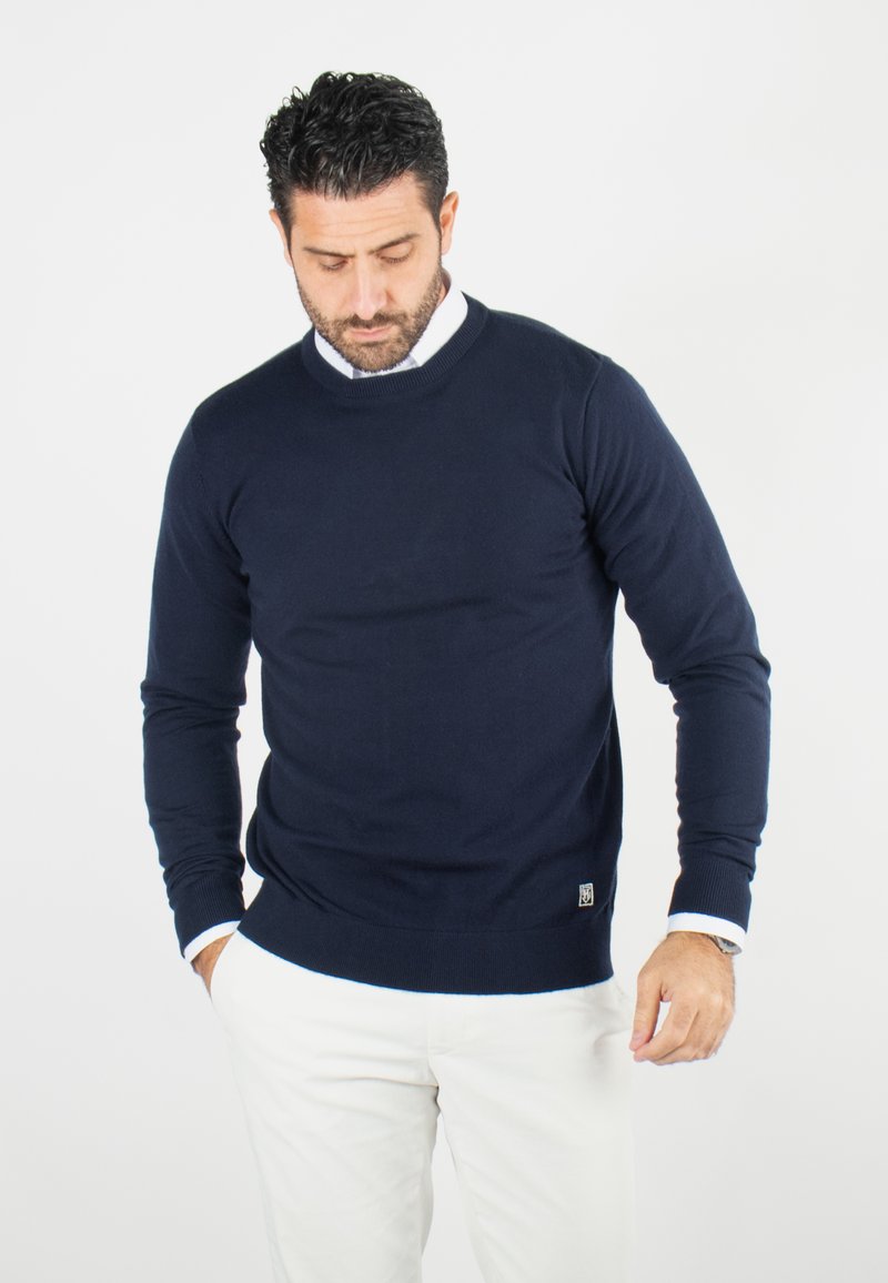 Pull en tricot bleu marine avec des manches longues, un col rond et des poignets côtelés. Présente un petit logo près de l'ourlet. Texture douce.