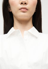 Γυναίκα με ίσια σκούρα μαλλιά, ντυμένη με λευκό πουκάμισο με κουμπιά, κοντινό πλάνο από το πηγούνι έως το πάνω μέρος του στήθους, με απλό φόντο.