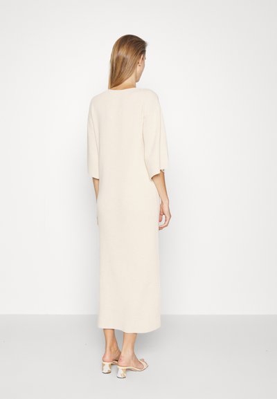 Μπεζ μακρύ φόρεμα με χαλαρή εφαρμογή, φαρδιά μανίκια, απαλή υφή και ίσια σιλουέτα, συνδυασμένο με ανοιχτόχρωμες σανδάλια.