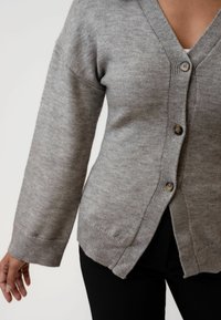 Šedý pletený cardigan s výstřihem do V, třemi knoflíky, žebrovaným lemem a postranními rozparky. Měkká textilie a uvolněný střih s dlouhými rukávy.