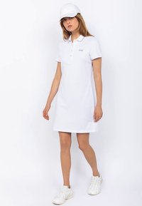 Vestido tipo polo blanco con mangas cortas, cuello con botones y corte recto. Se lleva con una gorra blanca a juego y zapatillas de color claro.