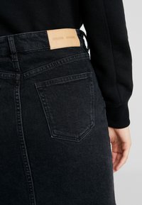 Jupe en denim noir à coupe droite, présentant une étiquette en cuir sur la ceinture et les poches arrière. La texture semble lisse et robuste.