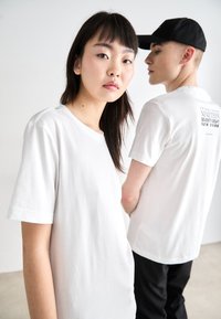 White cotton T-shirts with short sleeves, featuring a crew neckline; one shirt has a printed logo in black on the back. Black cap worn by one model.