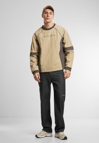 Beige and brown long-sleeve top with contrasting panels, featuring "Another Cotton" text. Gray jeans and beige sneakers complete the outfit.