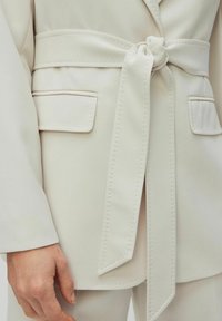 Světle béžový blazer s detailem vázaného pásku. Má hladkou látku, strukturovaný design a dvě ploché kapsy na přední straně.