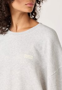 Svijetlosiva majica dugih rukava od mekane tkanine, s velikim krojem, zaobljenim izrezom i malim "American Vintage" logotipom u bijeloj boji.