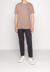 Camiseta de manga corta rosa clara con un pequeño logo amarillo en el pecho, combinada con pantalones oscuros y zapatillas blancas. Fondo liso.