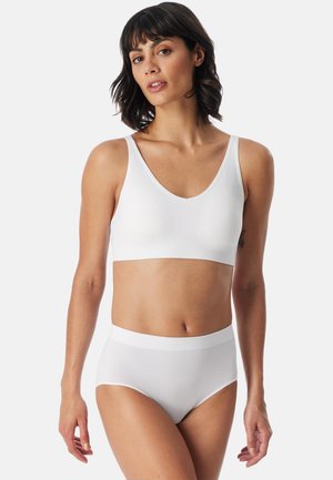 Kvinde med skulderlangt mørkt hår iført en hvid sømløs V-hals bh og høj taljebund, stående mod en ensfarvet baggrund.