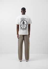 Bílá bavlněná tričko s černým grafickým potiskem na zádech, s kruhovým designem a textem. Kombinováno s béžovými cargo kalhotami.