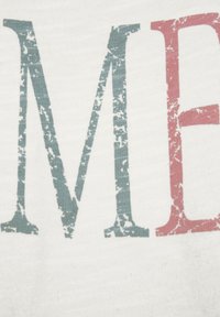 Witte stof met grote, versleten letters. De letter "M" in teal en "E" in rood, beide met een versleten textuur.