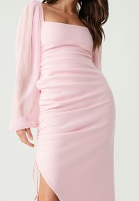 Woman wearing a light pink ruched midi dress with one long sheer sleeve and a square neckline, posing against a plain background.