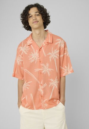 Jeune personne aux cheveux bouclés portant une chemise corail à manches courtes avec des motifs de palmiers blancs et un pantalon clair.