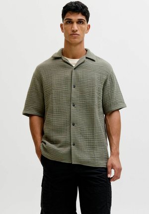 Uomo che indossa una camicia verde oliva a maniche corte con bottoni su una maglietta bianca e pantaloni neri, in piedi con le mani nelle tasche.
