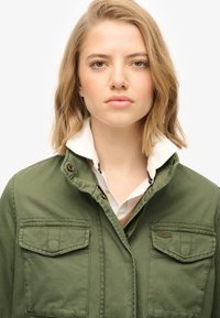 Olijfgroene jas met een gestructureerde kraag, voorzakken en een rits. Onder de jas is een witte shirtkraag zichtbaar. Soepele stoftextuur.