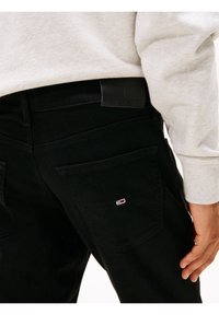 Mustat farkut, joissa on sileä pinta, nappikiinnitteiset takataskut ja vyötäröllä logoplaatti, pidetään vaaleanharmaan collegeneuleen alla.