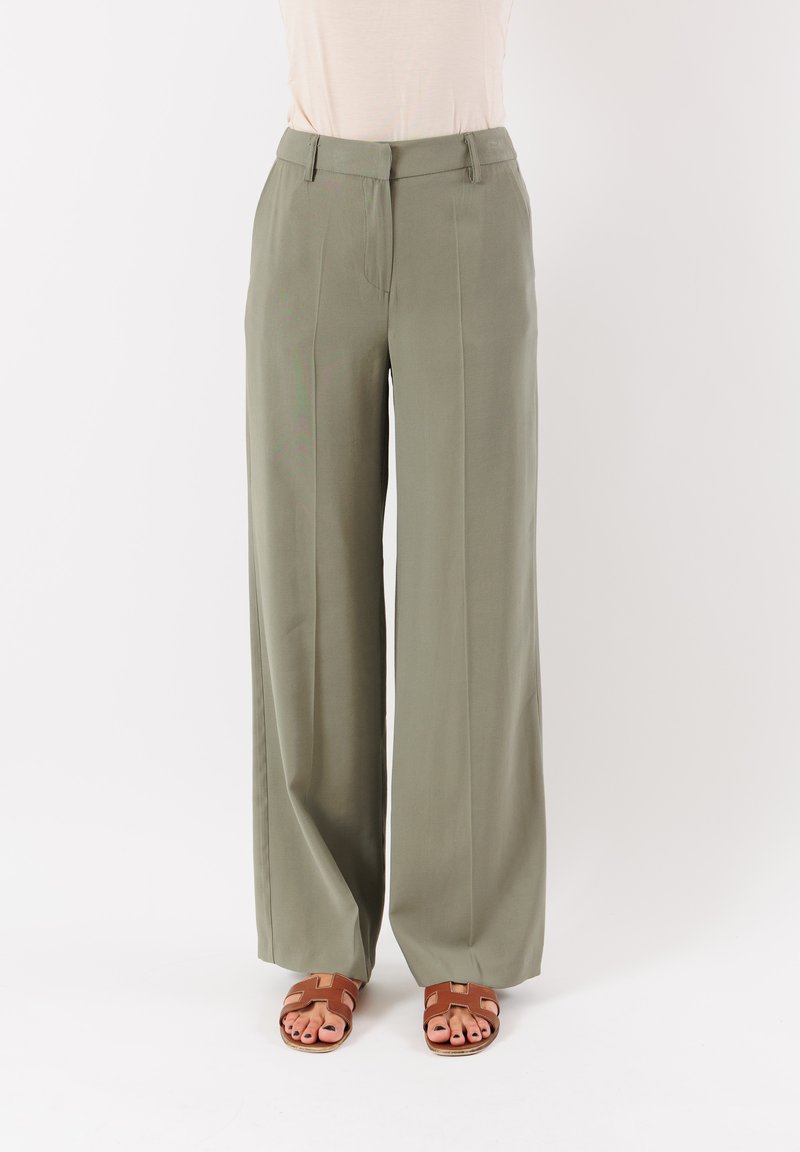 Pantaloni a gamba larga verde oliva realizzati in tessuto leggero, con pieghe frontali e vita sagomata. Indossati con sandali marroni.
