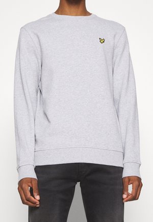 Mand iført en lysegrå langærmet crewneck-sweatshirt med lille gul fuglelogo på venstre bryst og sorte bukser.