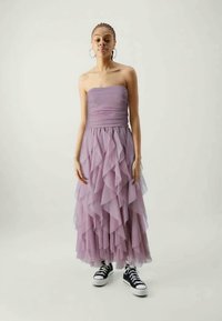 Φουσταριστό, strapless λιλά φόρεμα με επικαλυπτόμενες τulle ρυτίδες, φορεμένο με μαύρα αθλητικά sneakers, σε ουδέτερο φόντο.