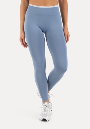 Kvinde iført lyseblå leggings med høj talje og hvide træningssko, stående med det ene ben krydset over det andet på en ensfarvet baggrund.