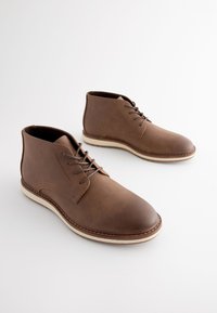 Stivaletti alla caviglia in pelle marrone con texture liscia, punta tonda e design con lacci. Presentano cuciture a contrasto e una suola bianca leggera.