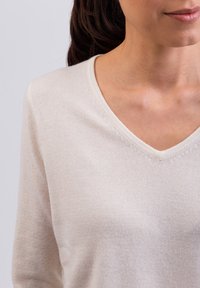 Sweter w serek w jasnym kremowym kolorze. Miękka tekstura, długie rękawy oraz subtelne detale przeszycia wzdłuż dekoltu.