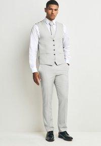 Costume gris clair avec un gilet ajusté et un pantalon sur mesure. Chemise blanche et cravate assortie. Chaussures habillées noires. Les boutons du gilet sont foncés.