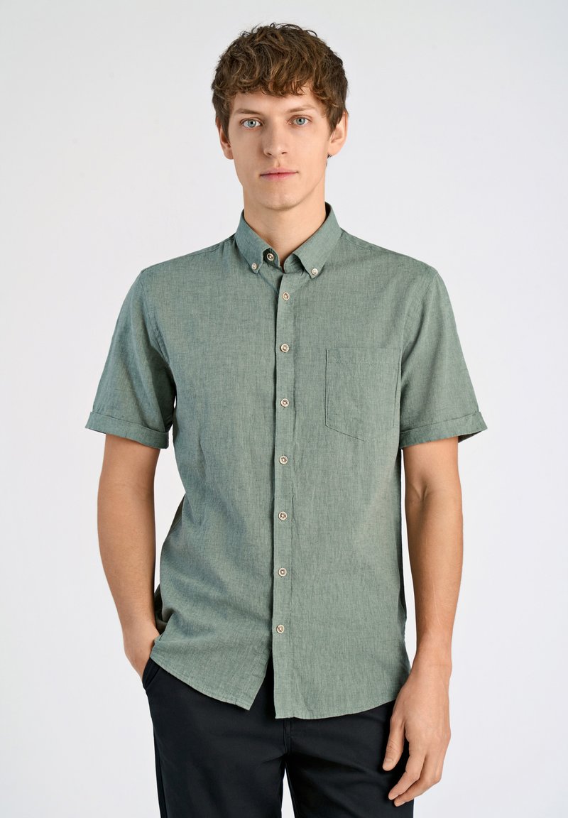Camisa de botones de manga corta en tejido verde texturizado, con un bolsillo en el pecho, un cuello abotonado y botones marrón claro.