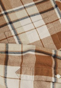 Bardasto in kremni karirasti material z tkano teksturo, ki ima modre in črne poudarne črte, eno žep in belo gumbno podrobnost.