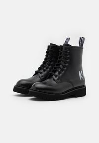 Botas de tobillo de cuero negro con punta redonda, diseño de cordones, suela gruesa negra y letras grises en el costado. Presentan tiradores de tela.