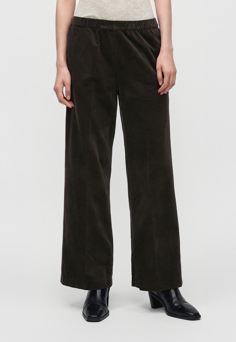 Personne portant un pantalon en velours côtelé marron foncé ample avec une taille élastique et des bottines noires à talons, debout devant un fond uni.