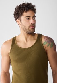 Khaki-grünes, geripptes Tanktop mit breiten Trägern, subtiler Textur und tailliertem Design, das ein florales Tattoo auf der Schulter zeigt.