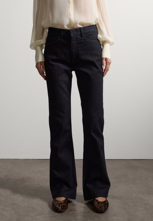 Person trägt schwarze High-Waist-Jeans, cremefarbene Bluse mit langen Puffärmeln und flache Schuhe mit Leopardenmuster, steht auf einem Betonboden.