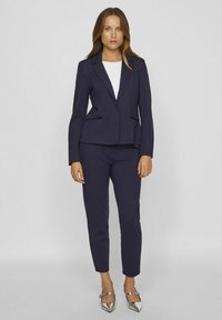 Mørkeblå tætsiddende blazer med matchende tapered bukser, der har en enkelt knaplukning, klapkrave og sidelommer. Glat stofstruktur.