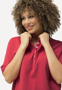 Polo shirt rosso a maniche corte con patta a bottoni e tessuto texturizzato. Il colletto è parzialmente aperto, rivelando una finitura liscia.