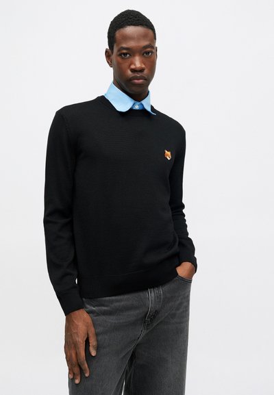 Mann iført svart genser med rever-emblem over blå skjorte og grå jeans, som står mot hvit bakgrunn.