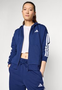 Mornaricno plavi dukserica s patentnim zatvaranjem i bijelim prugama na rukavima s Adidas logotipom. Uparen s odgovarajućim trenirkama koje imaju elastičnu traku u struku.