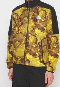 Giacca in pile con un motivo a tie-dye giallo e marrone, accenti neri sulle spalle e sulle maniche, chiusura con zip a tutta lunghezza e tasche laterali.