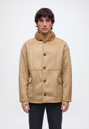 Un homme présente un manteau beige à col en peau lainée et à boutons foncés, ainsi qu’un pantalon, sur un fond clair uni.