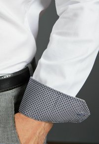Biała koszula z teksturowanej tkaniny; wzorzysty granatowy mankiet z małymi geometrycznymi kształtami; zbliżenie na koszulę i nadgarstek.