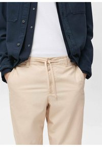 Béžové kalhoty na stažení se dvěma bočními kapsami, hladkou texturou a volným střihem, doplněné tmavě modrou košilí na knoflíky.